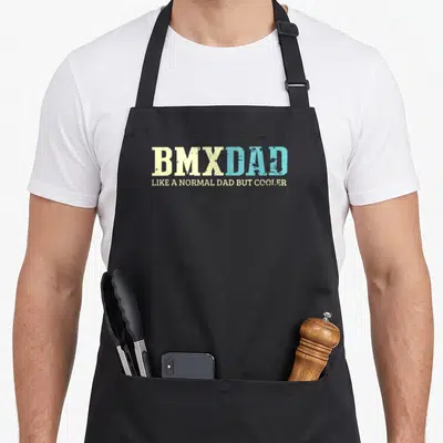 Normal bike apron - Stacked yellow and blue distressed typography reading BMXDAD LIKE A NORMAL DAD BUT COOLER — shown on a black cotton canvas apron against a white background