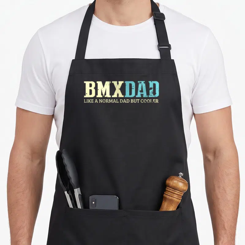 Normal bike apron - Stacked yellow and blue distressed typography reading BMXDAD LIKE A NORMAL DAD BUT COOLER — shown on a black cotton canvas apron against a white background