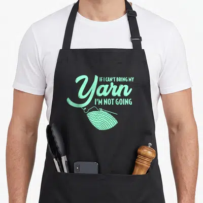 Crocheting knitting apron - Pastel green text reading If I Can't Bring My Yarn I'm Not Going above a yarn ball — shown on a black cotton canvas apron against a white background