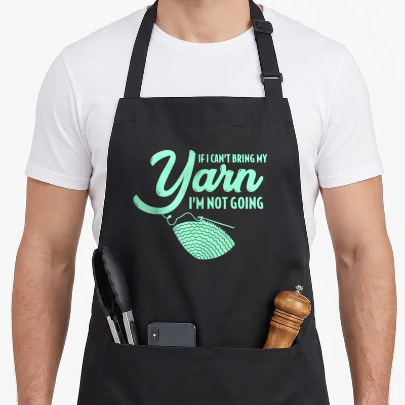 Crocheting knitting apron - Pastel green text reading If I Can't Bring My Yarn I'm Not Going above a yarn ball — shown on a black cotton canvas apron against a white background