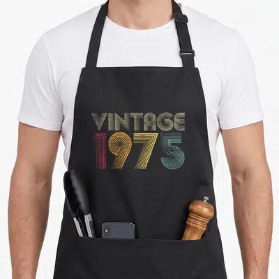 44th anniversary birthday apron - Retro VINTAGE 1975 text in beige, red, orange, yellow, and teal lines — shown on a black cotton canvas apron against a white background