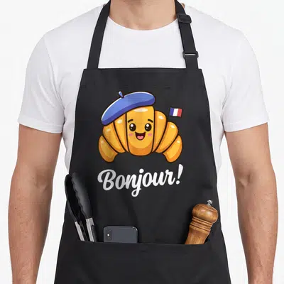 Croissant french apron - Cartoon orange croissant in blue beret holding a French flag with white Bonjour text below — shown on a black cotton canvas apron against a white background