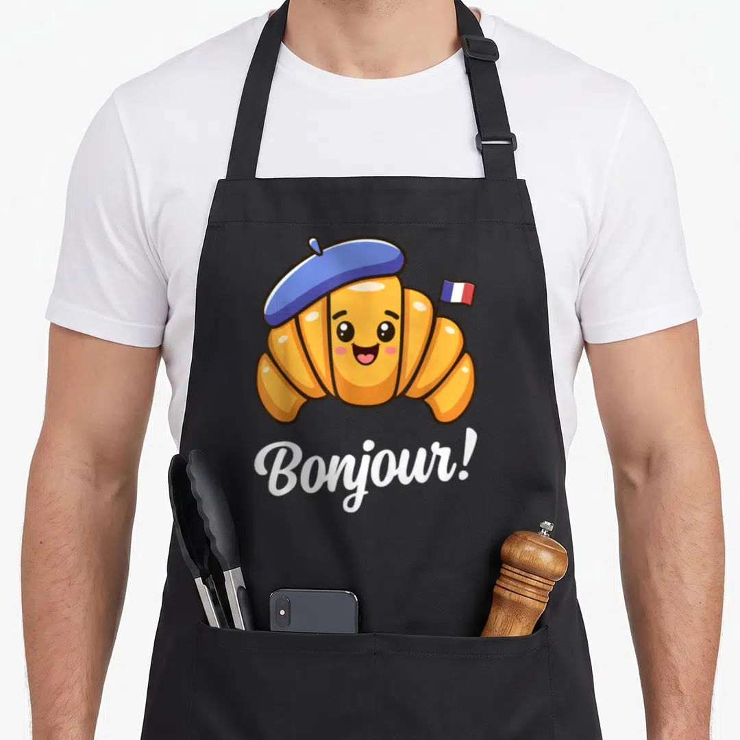 Croissant french apron - Cartoon orange croissant in blue beret holding a French flag with white Bonjour text below — shown on a black cotton canvas apron against a white background