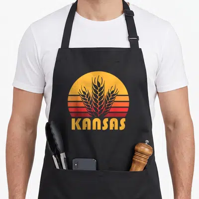 Kansas farming apron - Retro sunburst with yellow orange red grey stripes and grey wheat stalks above KANSAS text — shown on a black cotton canvas apron against a white background