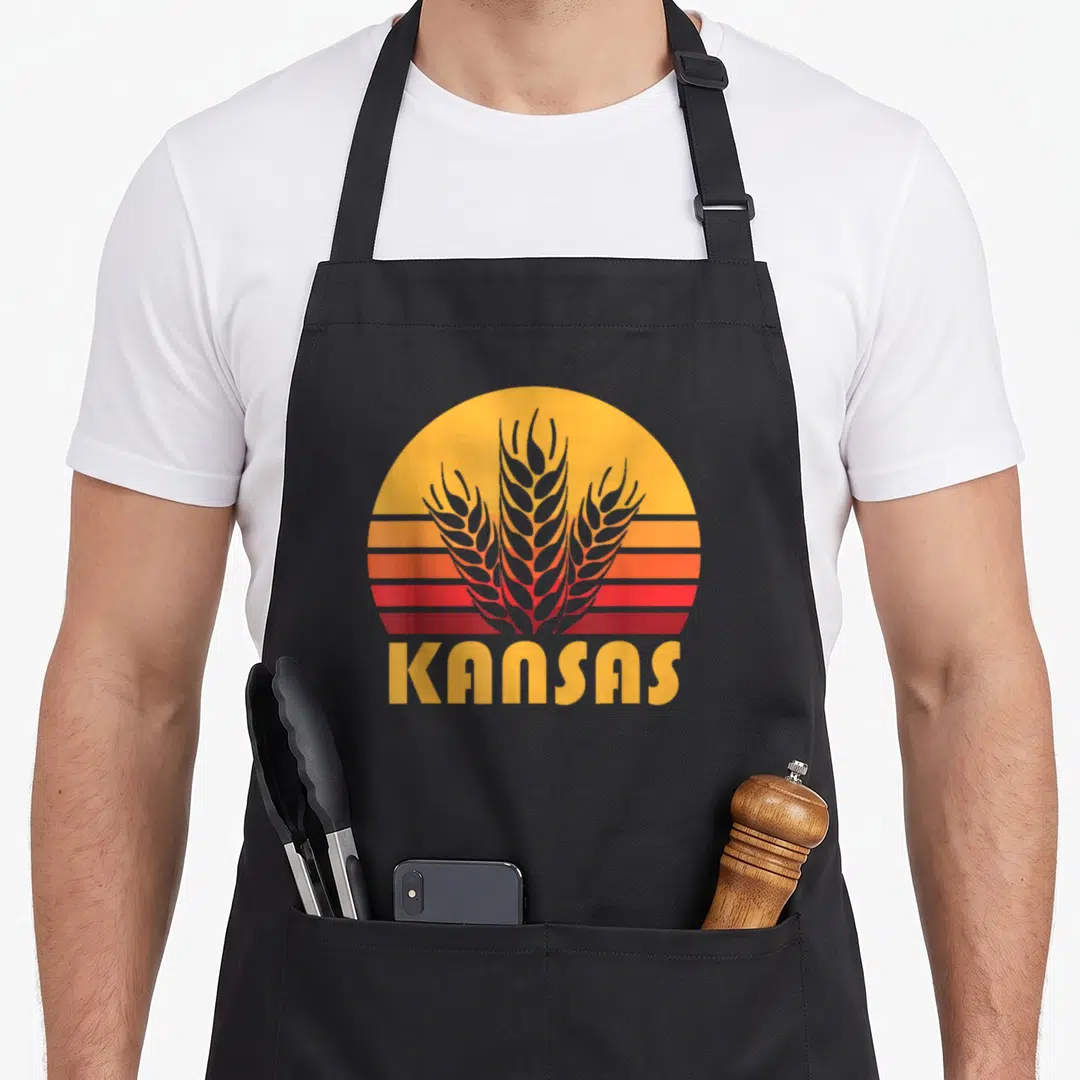 Kansas farming apron - Retro sunburst with yellow orange red grey stripes and grey wheat stalks above KANSAS text — shown on a black cotton canvas apron against a white background