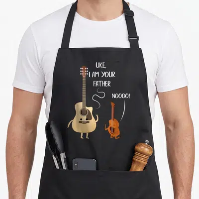 Father ukulele apron - Cartoon tan guitar and brown kneeling ukulele with white text UKE I AM YOUR FATHER NOOOO — shown on a black cotton canvas apron against a white background