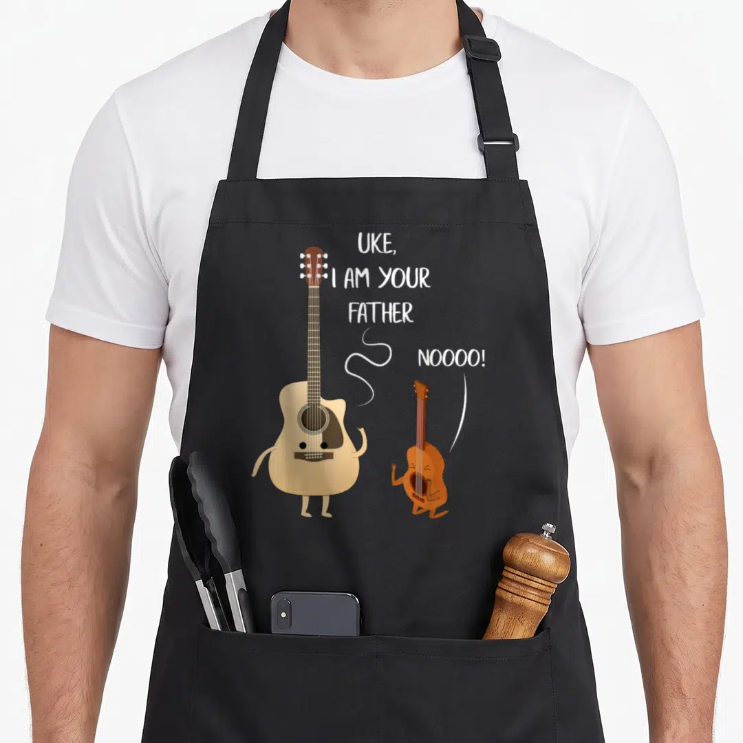 Father ukulele apron - Cartoon tan guitar and brown kneeling ukulele with white text UKE I AM YOUR FATHER NOOOO — shown on a black cotton canvas apron against a white background