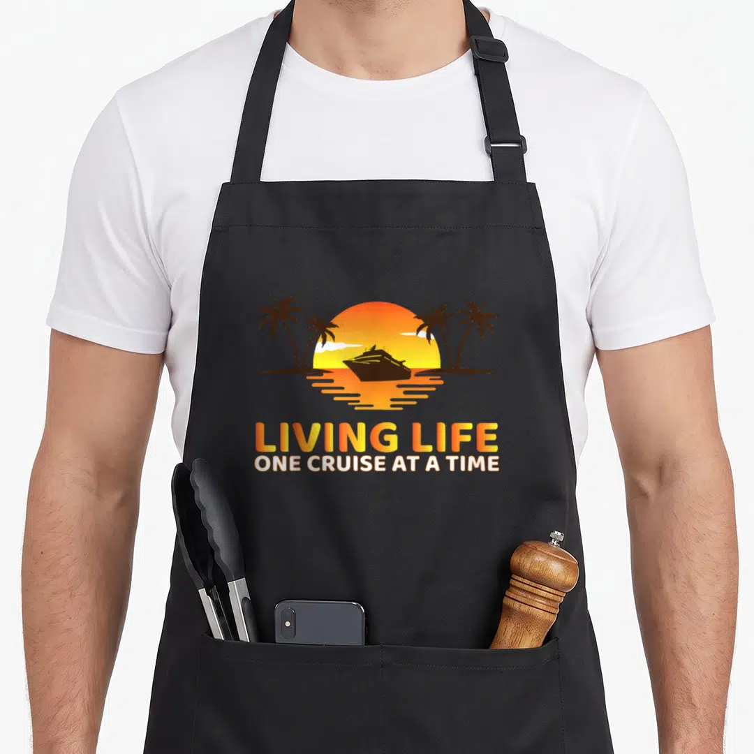 Living crazy cruise apron - Orange sunset with black cruise ship silhouette and stacked text between palm trees — shown on a black cotton canvas apron against a white background