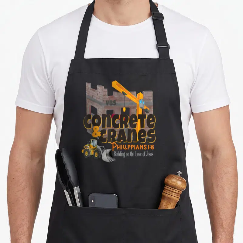 Crane building apron - Yellow crane, bulldozer, concrete block text, and Philippians 1:6 scripture on black fabric — shown on a black cotton canvas apron against a white background