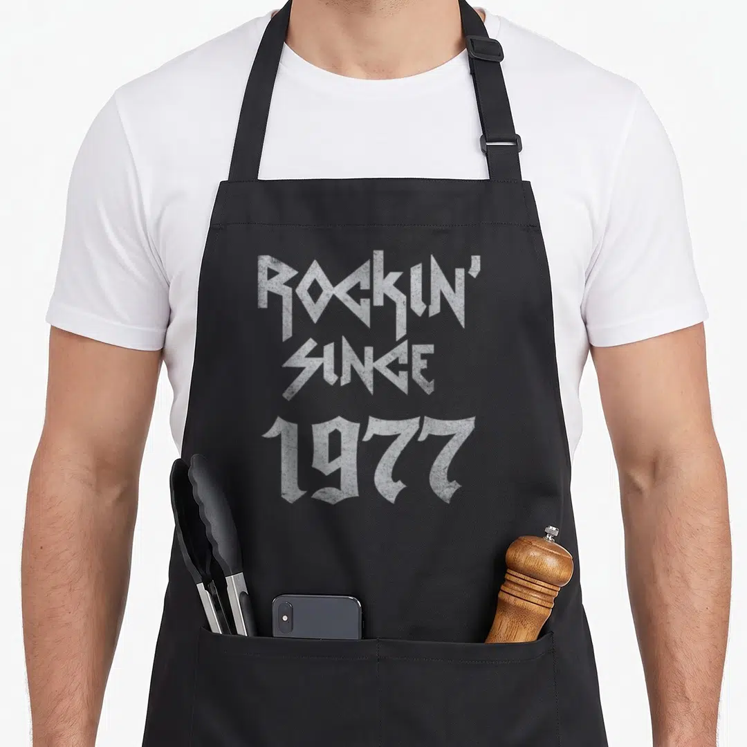Retro rockin birthday apron - White distressed typography reading ROCKIN SINCE 1977 stacked vertically — shown on a black cotton canvas apron against a white background