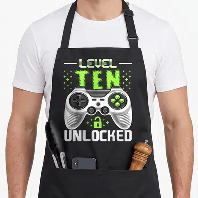 Level unlocked video gamer birthday apron - Game controller with LEVEL TEN UNLOCKED text and open green lock icon — shown on a black cotton canvas apron against a white background