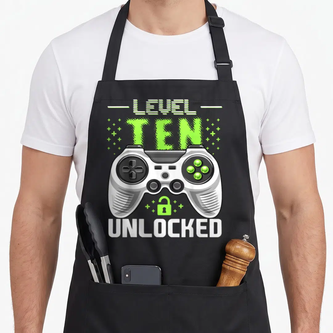 Level unlocked video gamer birthday apron - Game controller with LEVEL TEN UNLOCKED text and open green lock icon — shown on a black cotton canvas apron against a white background