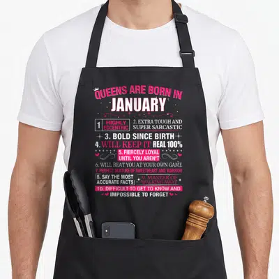 January queen apron - Queens Are Born In January text list with pink and white typography, crown, hearts and stars — shown on a black cotton canvas apron against a white background