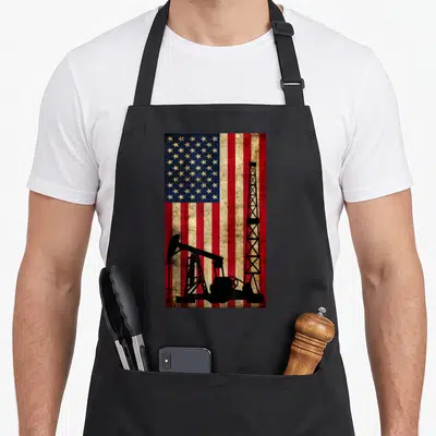 Landman oil mom apron - Distressed US flag with black oil derrick and pumpjack silhouettes — shown on a black cotton canvas apron against a white background
