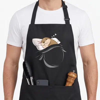 Pajamas animal apron - Cartoon otter lying on pillow under grey blanket with ZZZ text floating above — shown on a black cotton canvas apron against a white background
