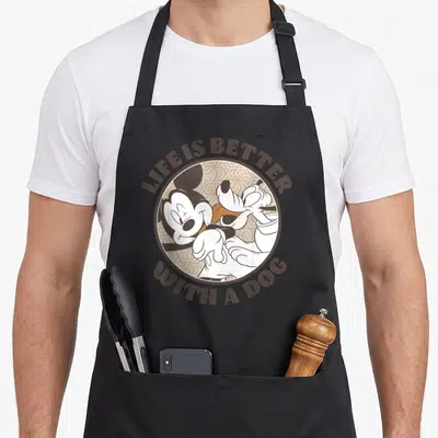 Better dog apron - Mickey and Pluto hugging inside circle with Life Is Better With A Dog text — shown on a black cotton canvas apron against a white background