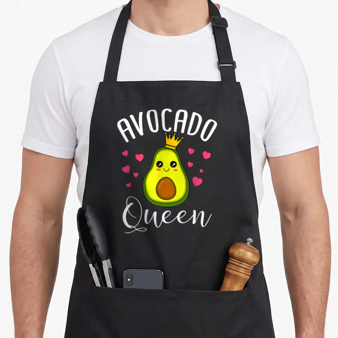 Queen avocado apron - Cartoon avocado wearing a yellow crown surrounded by pink hearts with white text — shown on a black cotton canvas apron against a white background