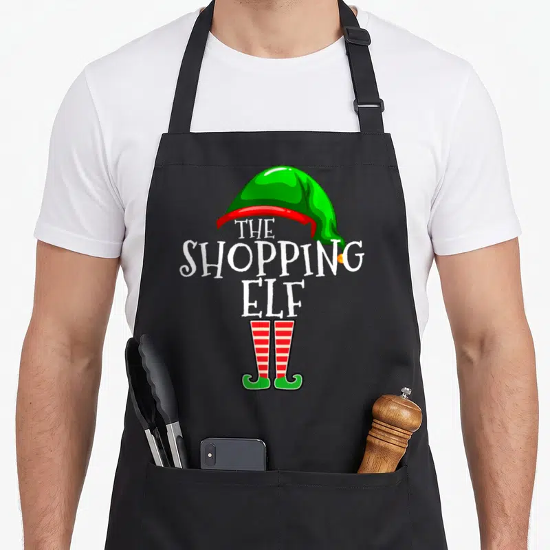 Shopping elf apron - Stacked white text THE SHOPPING ELF under green hat and red striped elf shoes — shown on a black cotton canvas apron against a white background