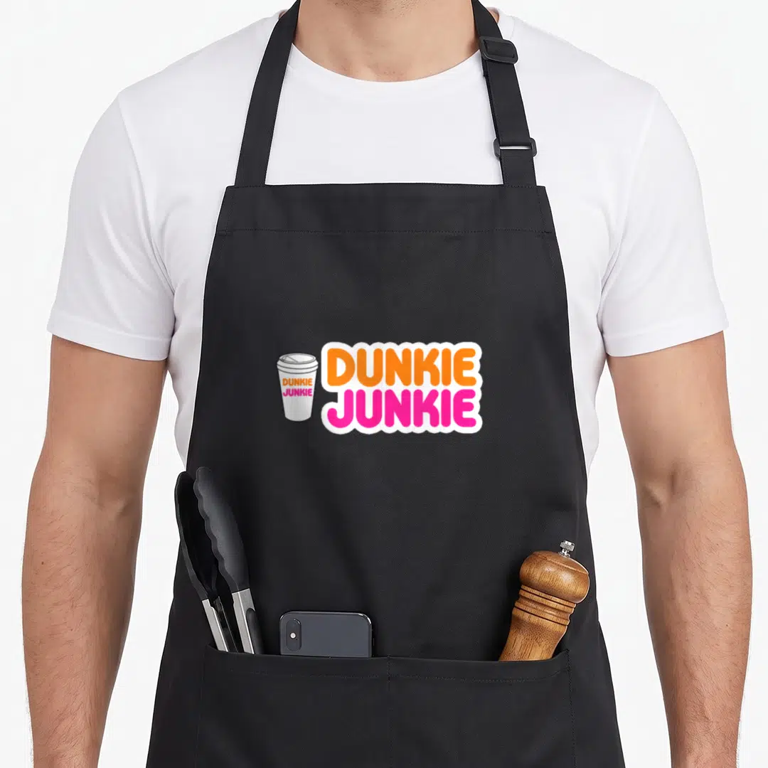 Dunkie coffee apron - Cartoon coffee cup with stacked orange and pink DUNKIE JUNKIE text in white bubble border — shown on a black cotton canvas apron against a white background