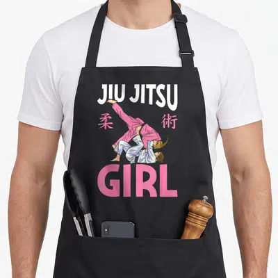 Martial bjj apron - JIU JITSU GIRL text with two grapplers in pink and blue gis flanked by pink kanji — shown on a black cotton canvas apron against a white background