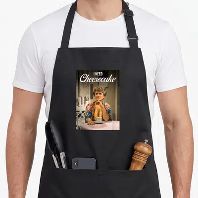 Blanche golden apron - Photo of woman at pink table with text I NEED Cheesecake in white script and block letters — shown on a black cotton canvas apron against a white background