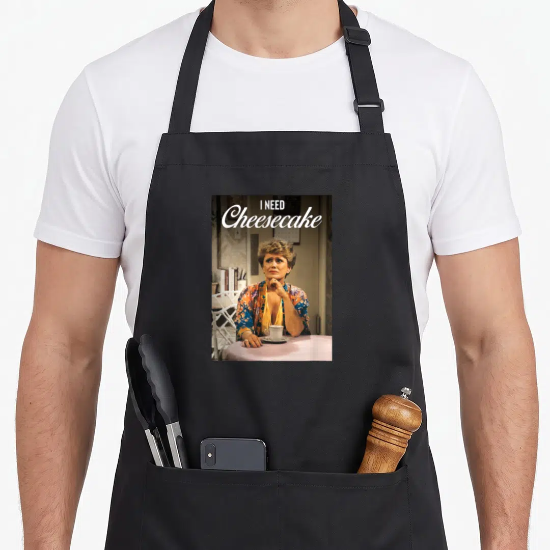Blanche golden apron - Photo of woman at pink table with text I NEED Cheesecake in white script and block letters — shown on a black cotton canvas apron against a white background