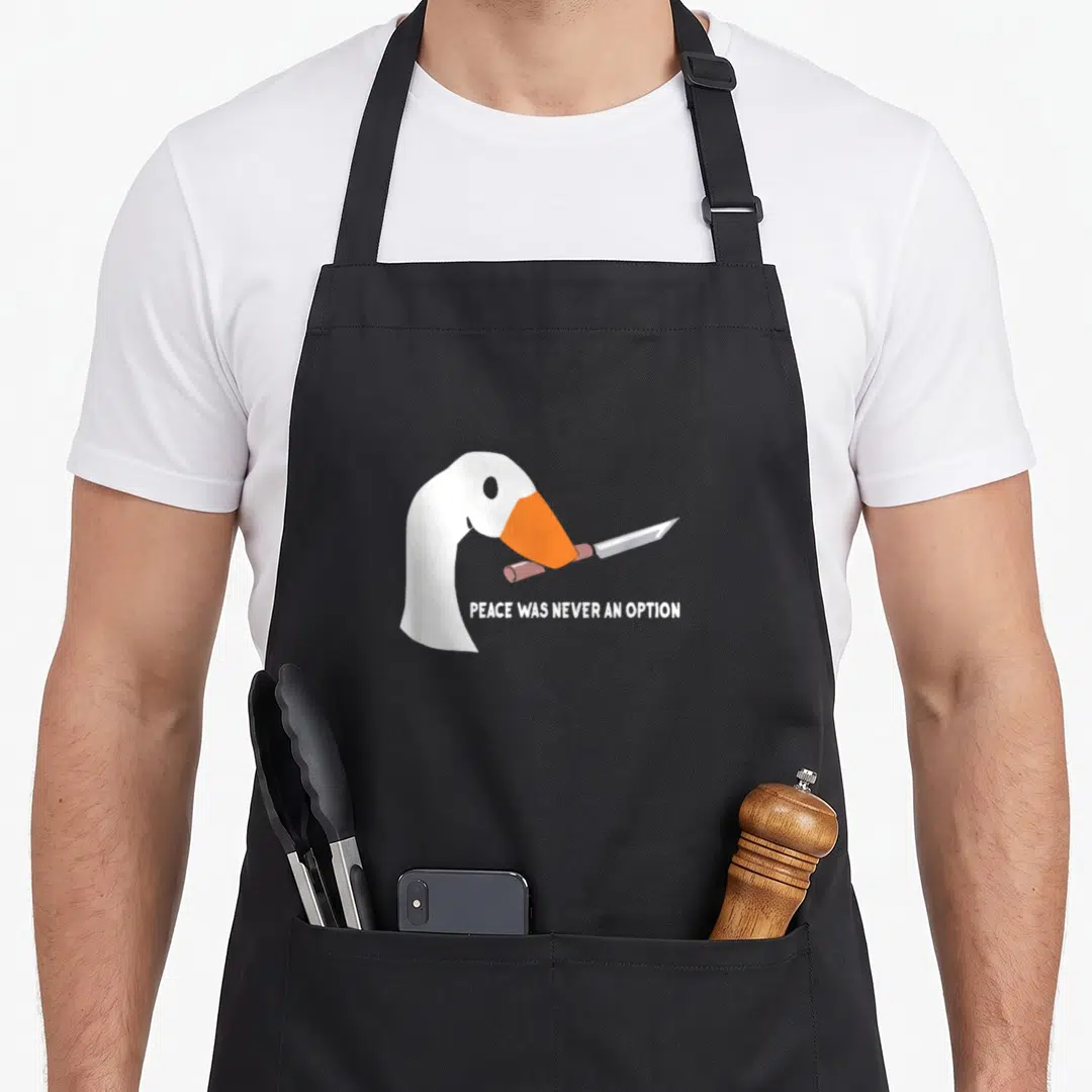 Funny peace goose apron - Cartoon white goose head holding a grey knife with brown handle and text below — shown on a black cotton canvas apron against a white background