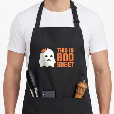 Ghost boo sheet halloween apron - White cartoon ghost with orange bow beside orange THIS IS BOO SHEET text — shown on a black cotton canvas apron against a white background
