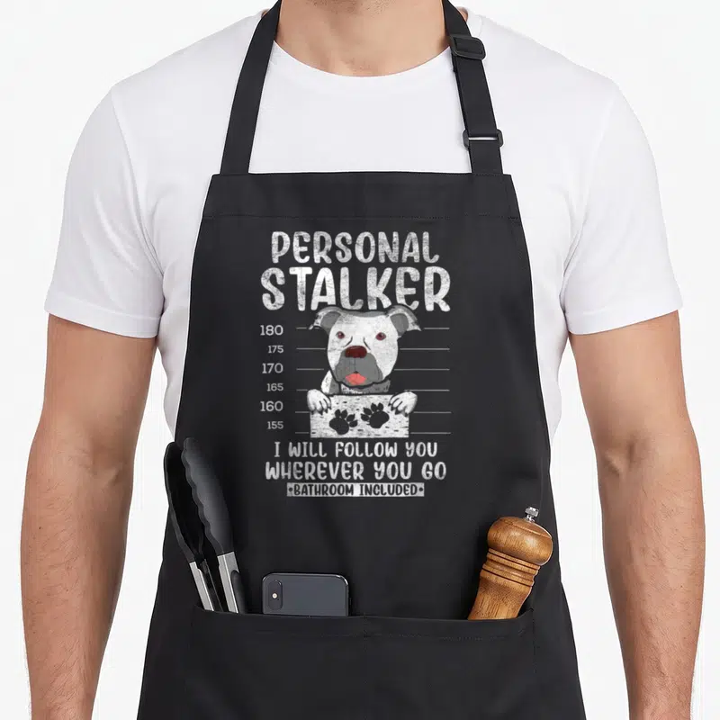 Stalker personal printed aprons - White dog peeking over height chart with personal stalker text — shown on a black cotton canvas apron against a white background