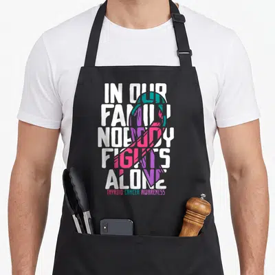 Family thyroid apron - White text IN OUR FAMILY NOBODY FIGHTS ALONE over teal pink purple ribbon with small bottom — shown on a black cotton canvas apron against a white background