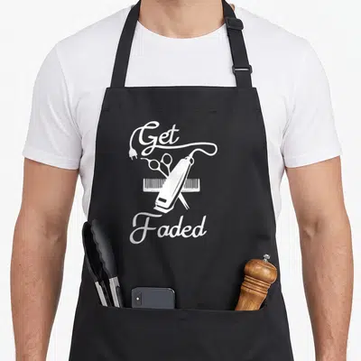 Faded haircut apron - White line art of hair clipper, scissors, comb and Get Faded text — shown on a black cotton canvas apron against a white background