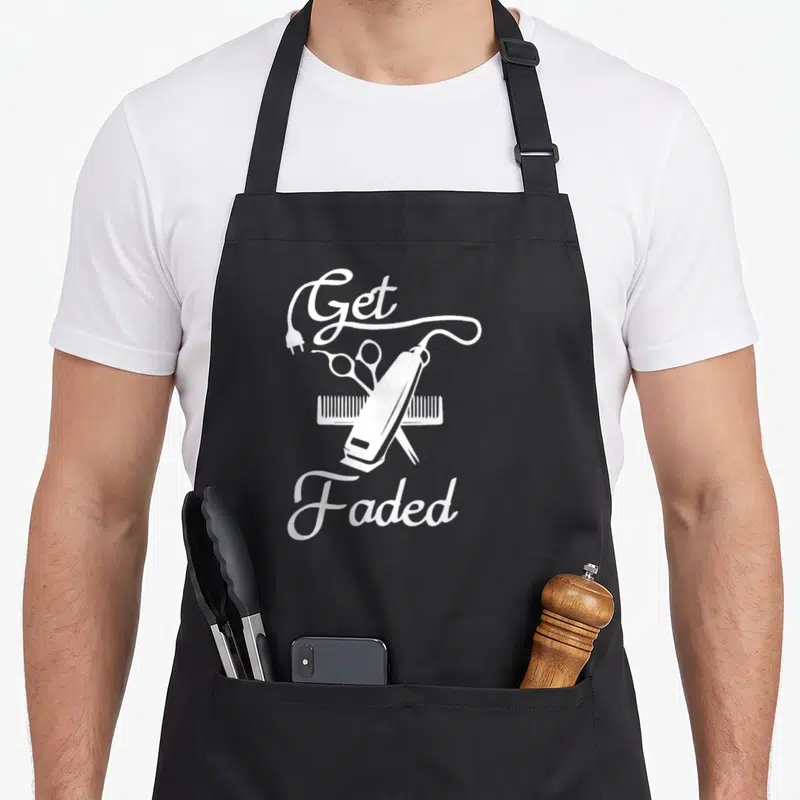 Faded haircut apron - White line art of hair clipper, scissors, comb and Get Faded text — shown on a black cotton canvas apron against a white background