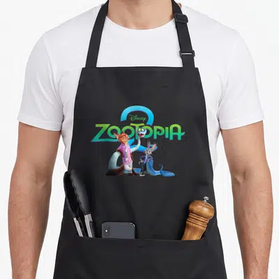 Gary novelty apron - Disney ZOOTOPIA logo with fox, rabbit, and blue snake in green and blue cartoon art — shown on a black cotton canvas apron against a white background