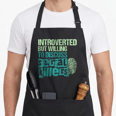 Introverted crime apron - Stacked green and teal text reading introverted willing discuss serial killers beside — shown on a black cotton canvas apron against a white background