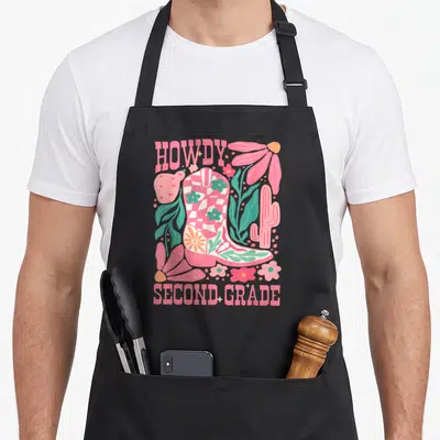 Howdy 2nd apron - Pink cowboy boot with flowers and HOWDY SECOND GRADE text in western cartoon style — shown on a black cotton canvas apron against a white background