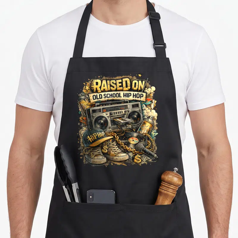 Street hip apron - Boombox, cap, sneakers, and raised on old school hip hop text in cartoon style — shown on a black cotton canvas apron against a white background