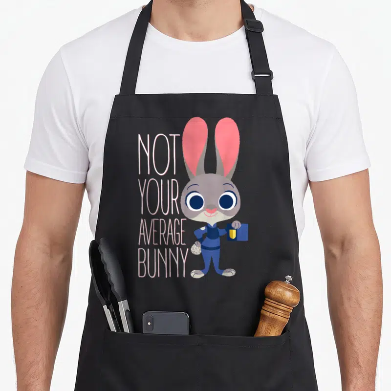 Judy bunny apron - Cartoon grey bunny in blue uniform holding a card next to stacked text reading Not Your Average Bunny — shown on a black cotton canvas apron against a white background