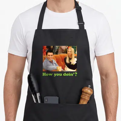 Friend unique apron - Joey and Phoebe photo on orange couch above lime green How you doin text — shown on a black cotton canvas apron against a white background