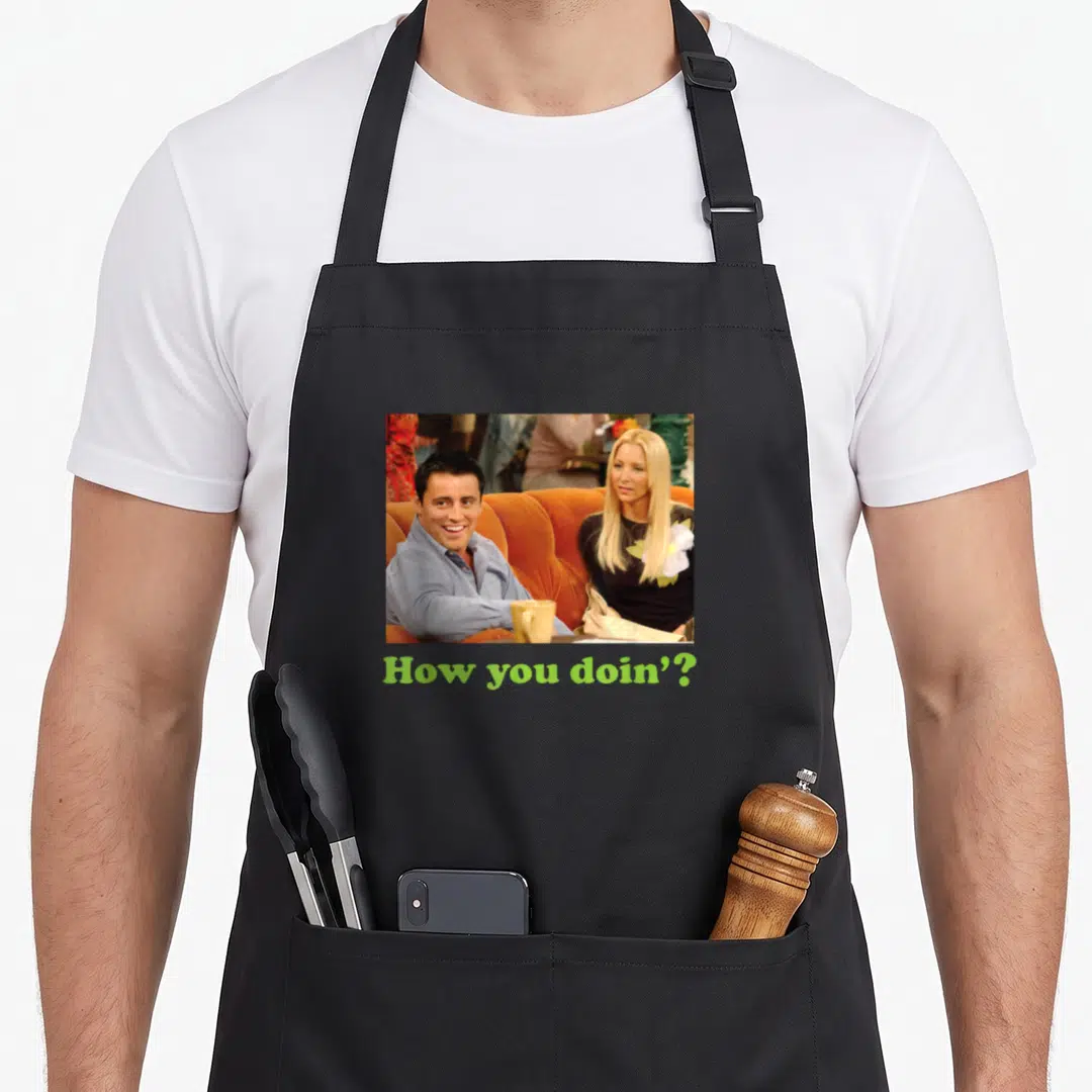 Friend unique apron - Joey and Phoebe photo on orange couch above lime green How you doin text — shown on a black cotton canvas apron against a white background