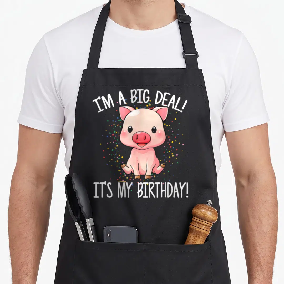 Deal birthday apron - Pink cartoon pig sitting with I'M A BIG DEAL! text and scattered confetti — shown on a black cotton canvas apron against a white background