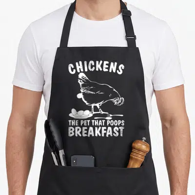 Poop chicken apron - Distressed white chicken illustration above eggs on a plate with fork and spoon and stacked text — shown on a black cotton canvas apron against a white background
