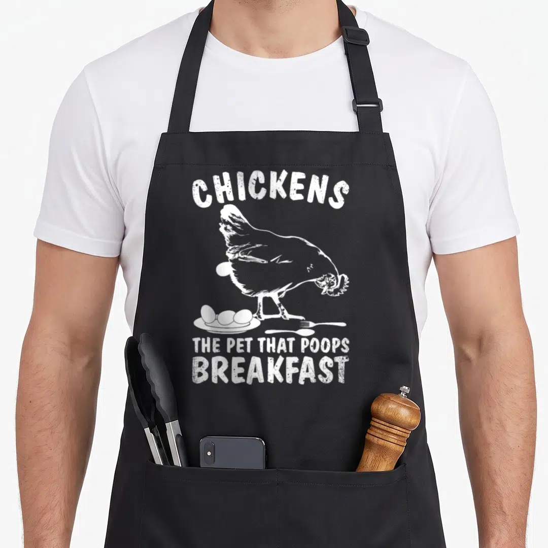 Poop chicken apron - Distressed white chicken illustration above eggs on a plate with fork and spoon and stacked text — shown on a black cotton canvas apron against a white background