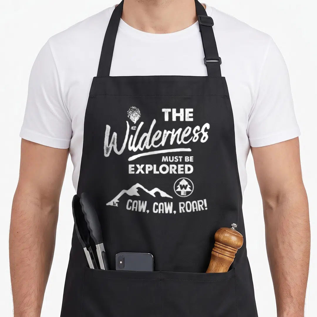 Wilderness camping apron - Distressed white text reading The Wilderness Must Be Explored with mountain and tree icons — shown on a black cotton canvas apron against a white background