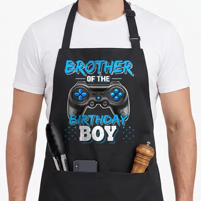 Brother birthday video game apron - Blue and white text surrounding a black game controller illustration on a grey — shown on a black cotton canvas apron against a white background