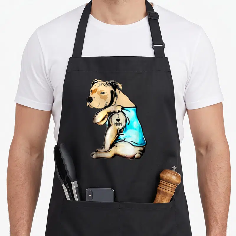 Pit tattoo apron - Tan dog in blue shirt with I heart MOM tattoo text on arm — shown on a black cotton canvas apron against a white background