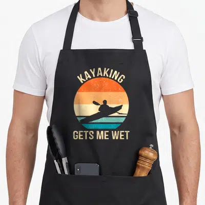 Kayaker wet aprons - Retro striped sun with kayaker silhouette and text KAYAKING GETS ME WET — shown on a black cotton canvas apron against a white background