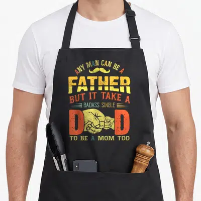 Father badass apron - Stacked text with yellow mustache and fist replacing letter D in red and yellow distressed font — shown on a black cotton canvas apron against a white background