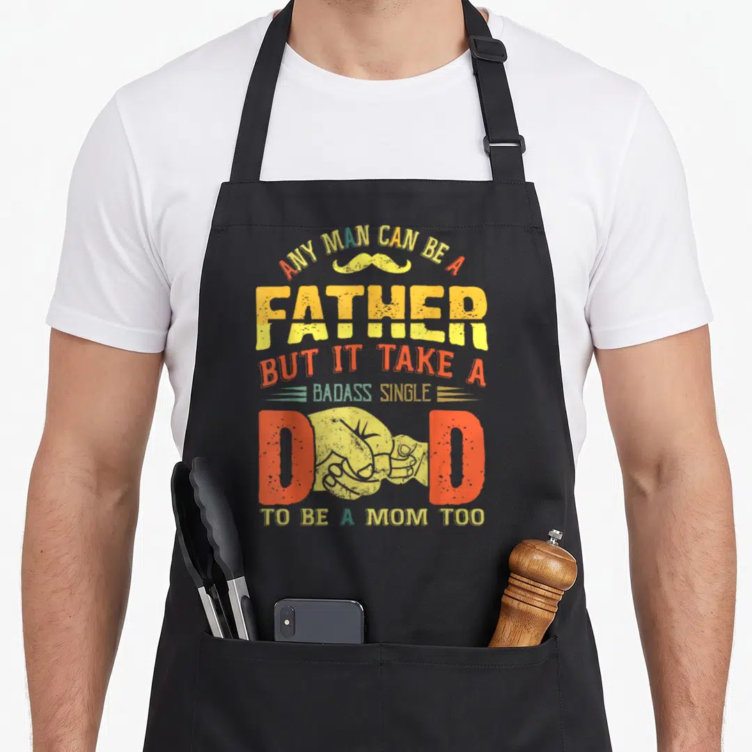 Father badass apron - Stacked text with yellow mustache and fist replacing letter D in red and yellow distressed font — shown on a black cotton canvas apron against a white background