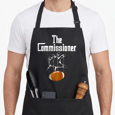 Commissioner football apron - White text The Commissioner above a hand puppeteering an orange football with strings — shown on a black cotton canvas apron against a white background