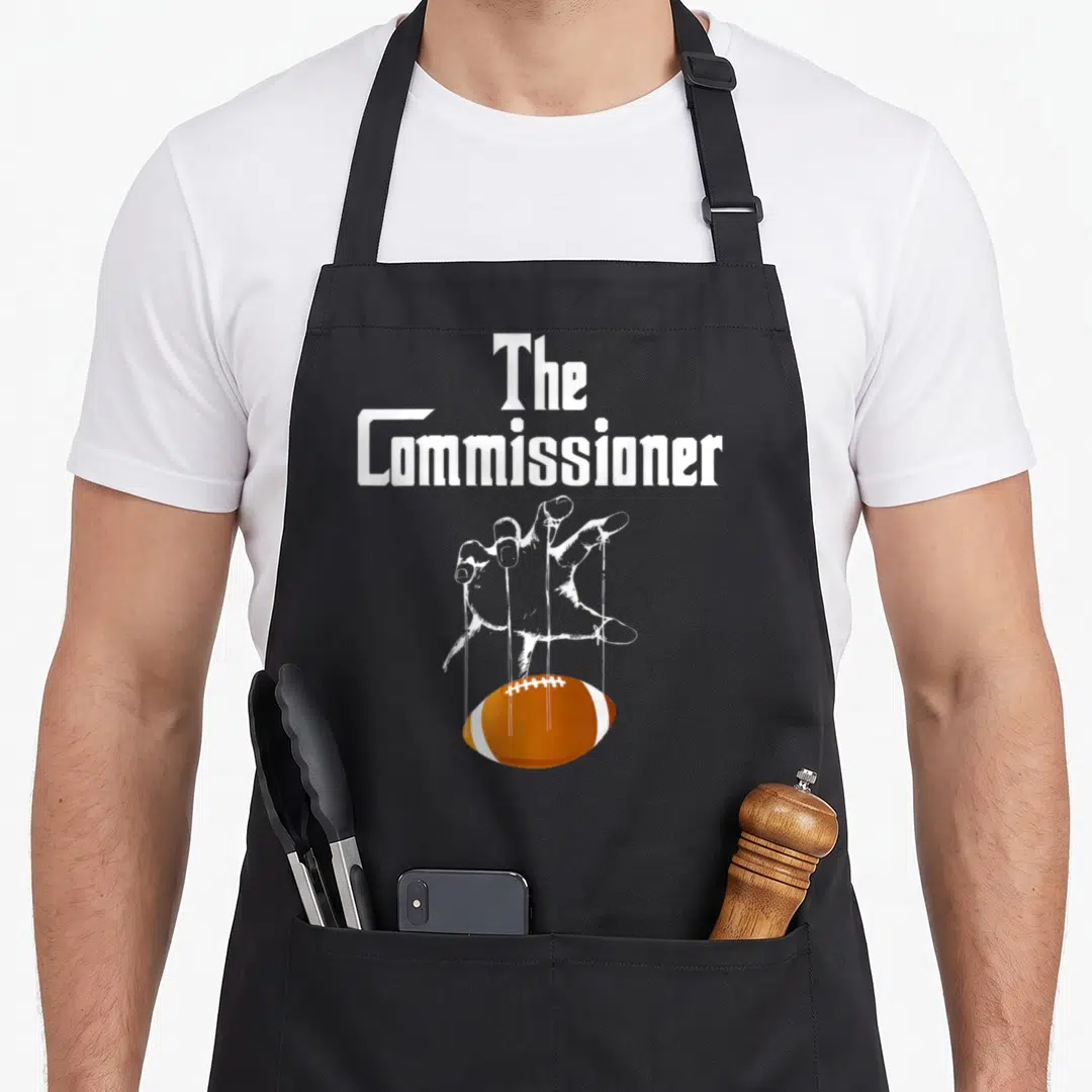 Commissioner football apron - White text The Commissioner above a hand puppeteering an orange football with strings — shown on a black cotton canvas apron against a white background